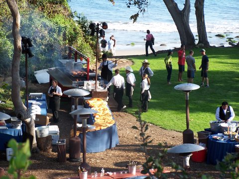 serving tables and grill set up next to a green lawn overlooking the beach and waves in the ocean